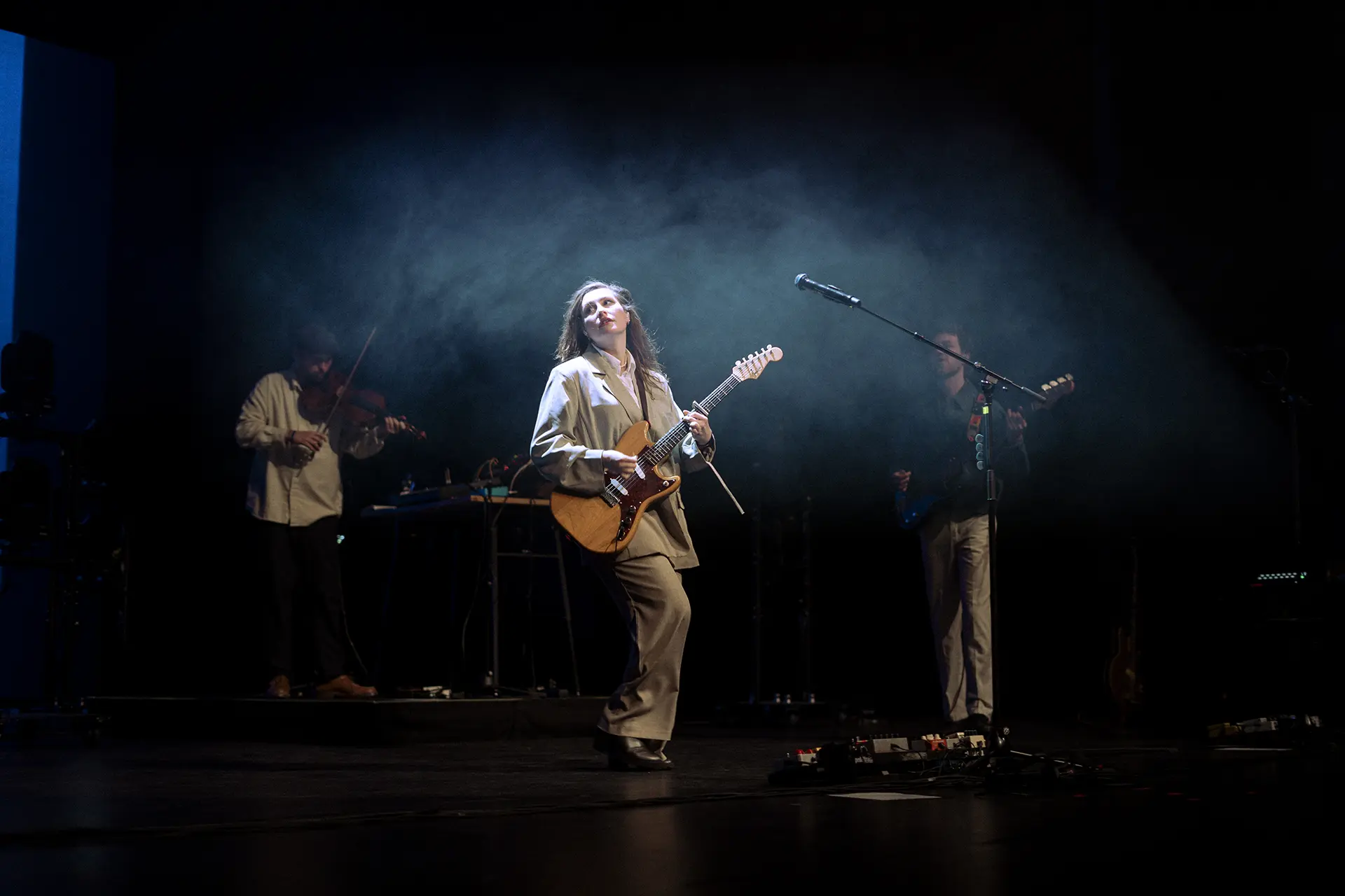 Een muzikante met een gitaar staat centraal op het podium terwijl zij wordt omringd door andere muzikanten in een sfeervolle, licht rokerige setting.