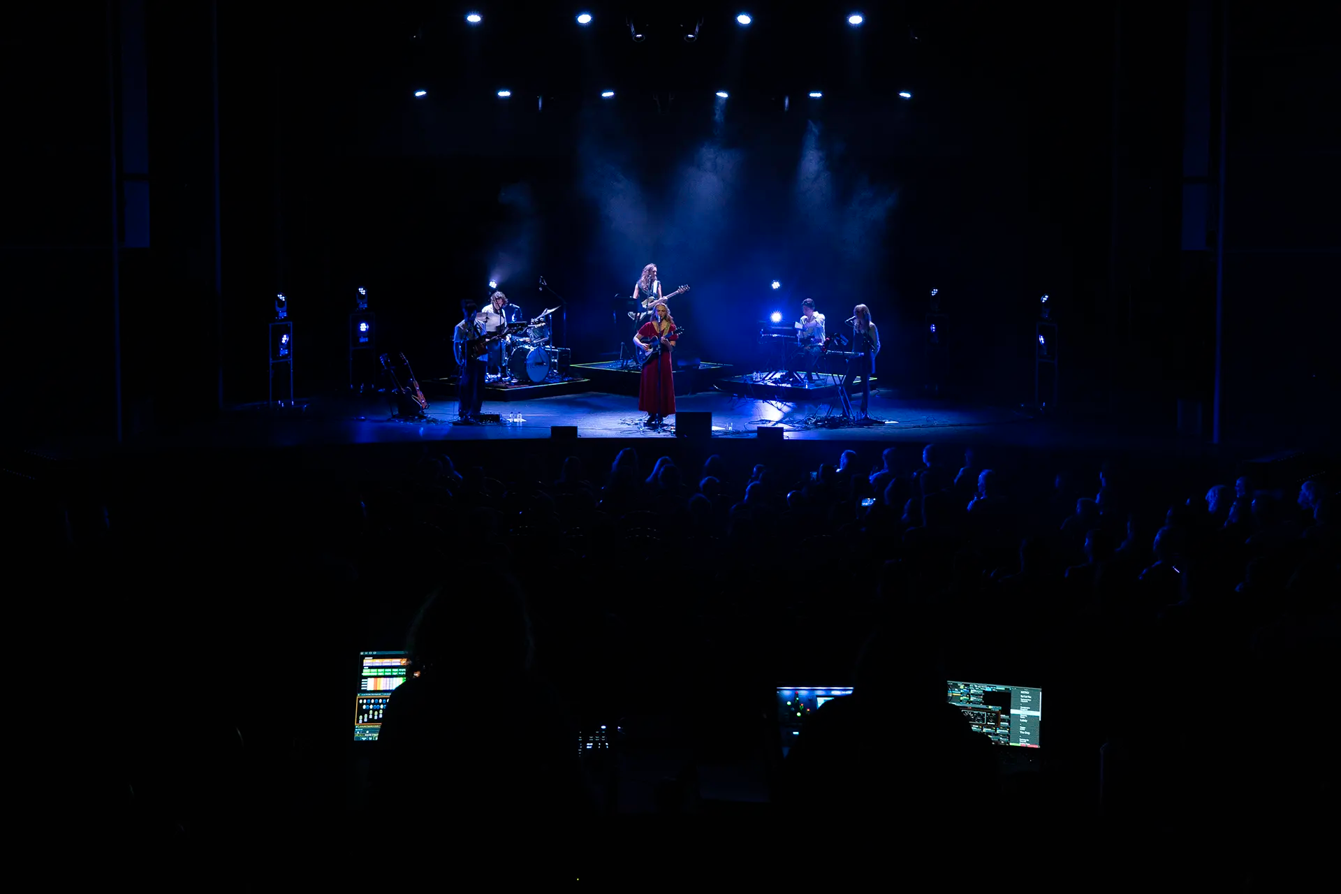 Een band speelt live op een podium verlicht door blauwe lichten, terwijl het publiek in de schaduw toekijkt.
