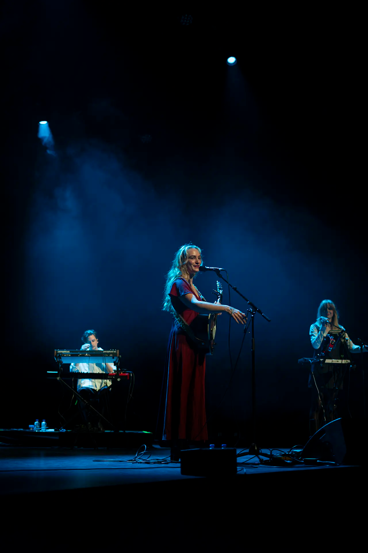 Een zangeres met een gitaar staat in het middelpunt van het podium terwijl twee muzikanten haar begeleiden, omringd door mysterieus blauw licht.