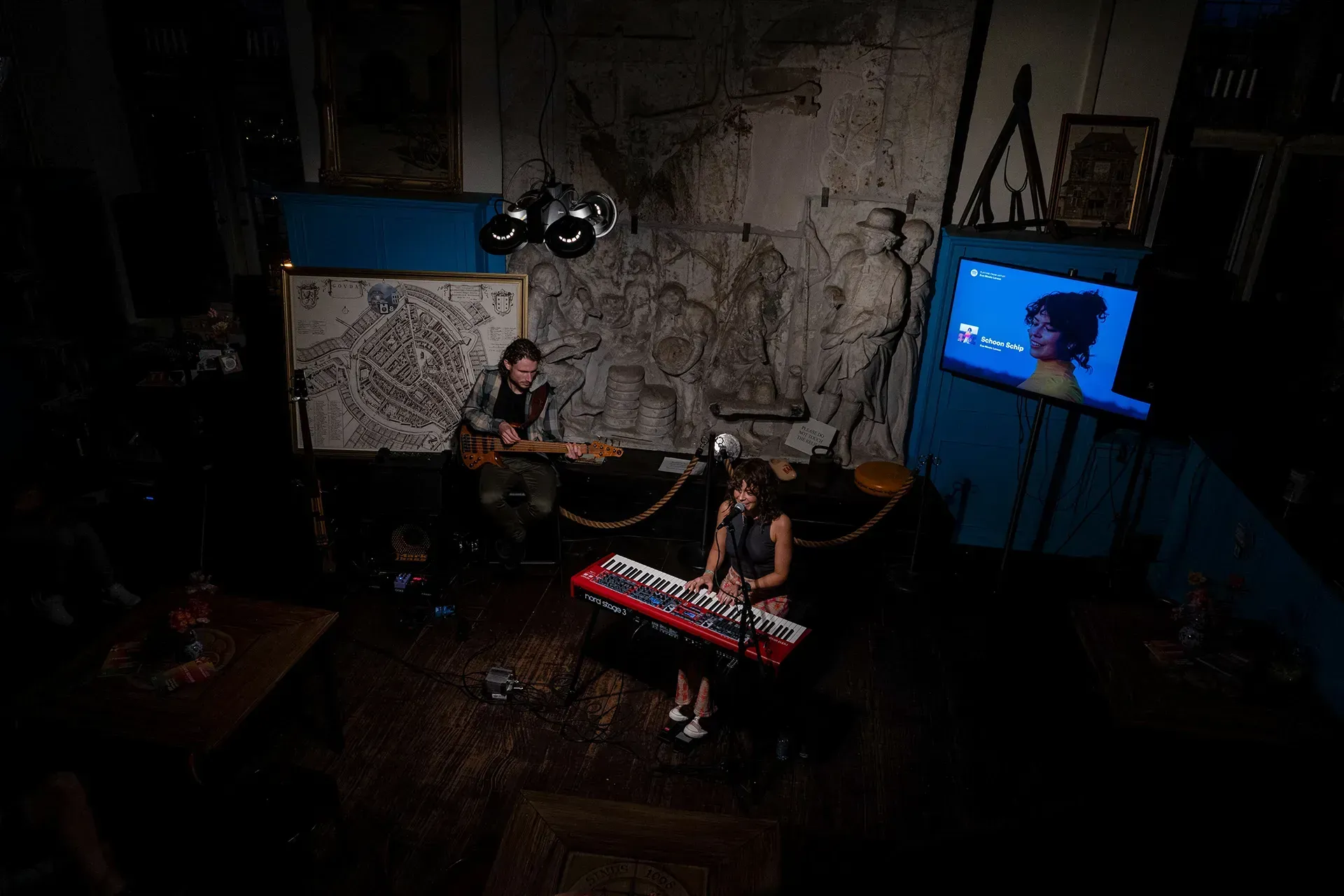 Een muzikante speelt op een rode keyboard terwijl een gitarist op de achtergrond speelt, omringd door kunst en een scherm met informatie.