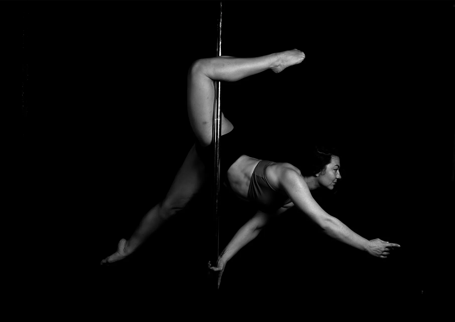 Een acrobate pose op een paal door een vrouw, vastgelegd in zwart-wit tegen een donkere achtergrond.
